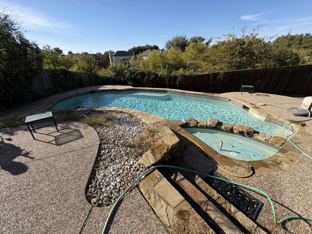 Outdoor swimming pool and attached hot tub in a backyard, surrounded by stone and concrete patio, with a garden hose on the ground—perfect for relaxing, but keep an eye out if your pool is losing water among the trees in the background.