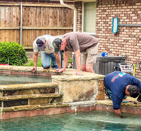 Three men kneel at the edge of a pool, inspecting and working on the water and pool structure beside a brick house, demonstrating expert pool leak detection in Dallas.