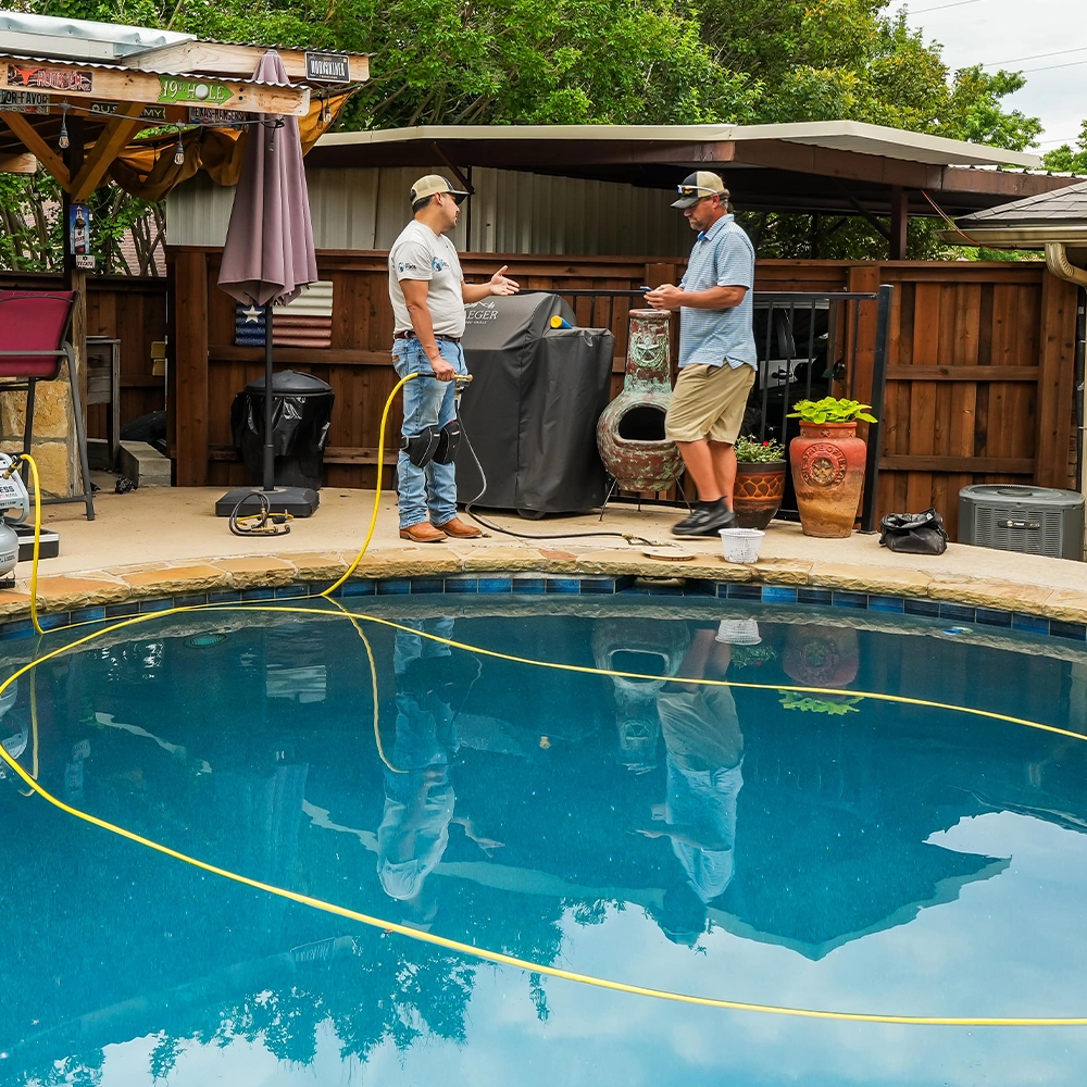 Two men stand beside a backyard swimming pool, discussing Pool Leak Detection in Prosper TX while holding tools and yellow cables; a covered patio and barbecue grill are in the background.