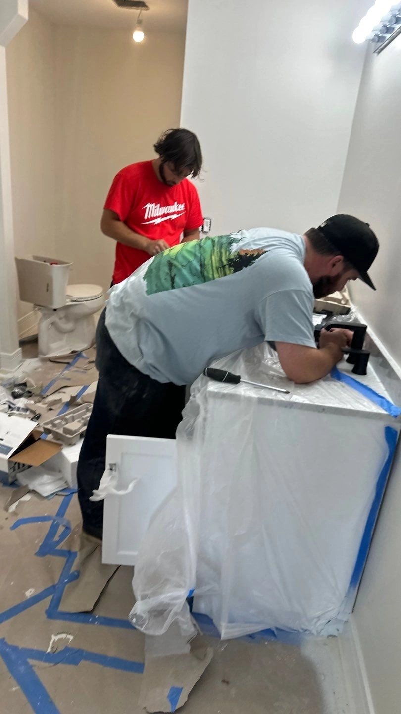 Two workers renovating a bathroom, one installing a sink, the other painting.