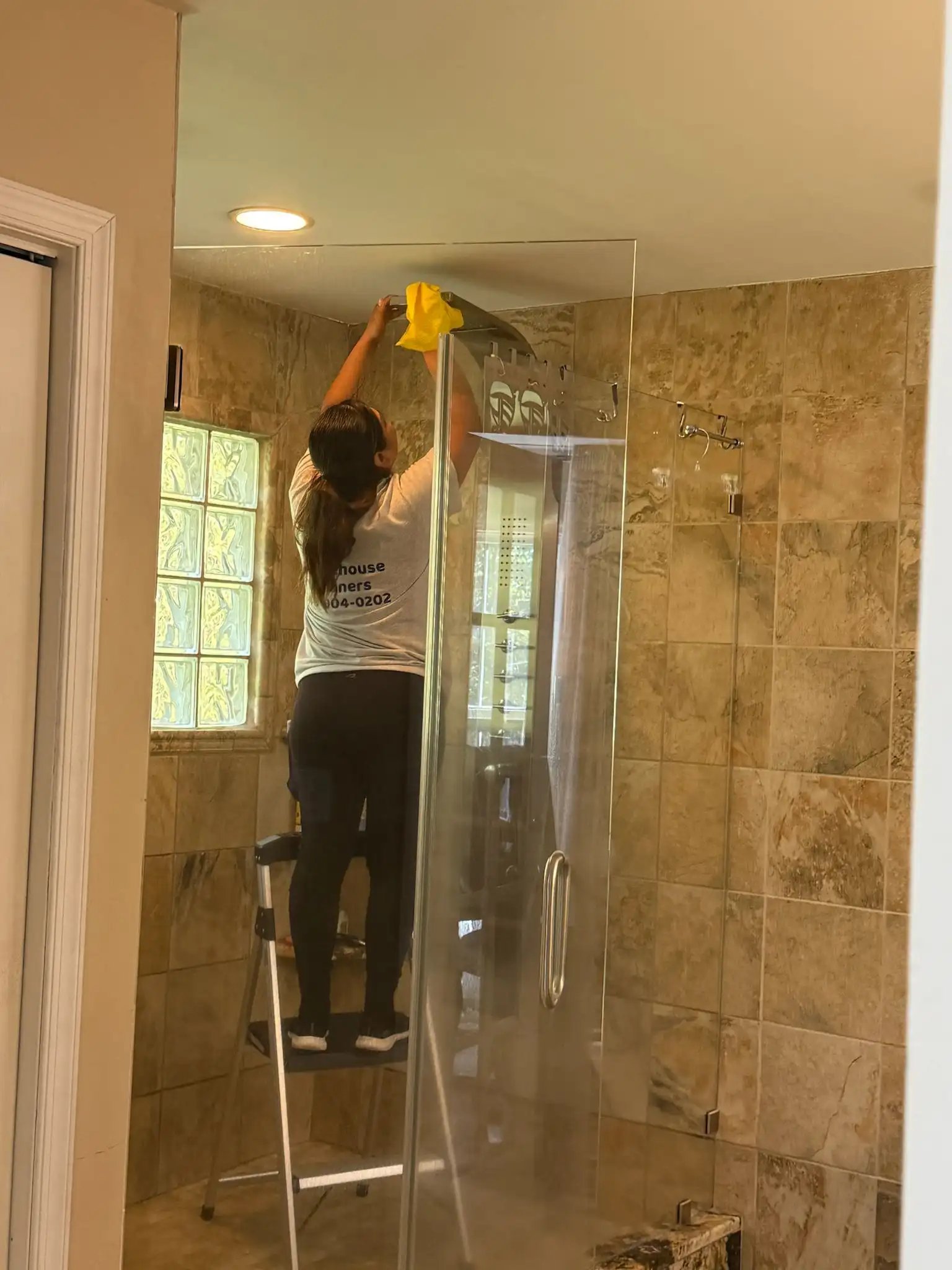 Person cleaning a glass shower enclosure on a ladder