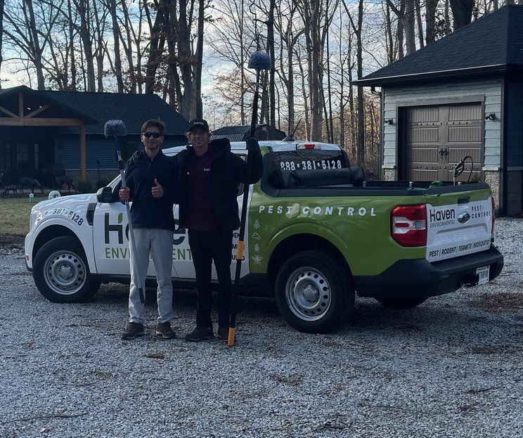 Two pest control professionals stand by a truck in a wooded area.