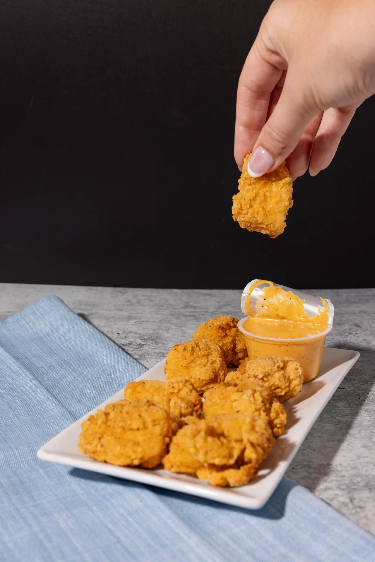 Hand holding chicken nugget above a plate with dipping sauce