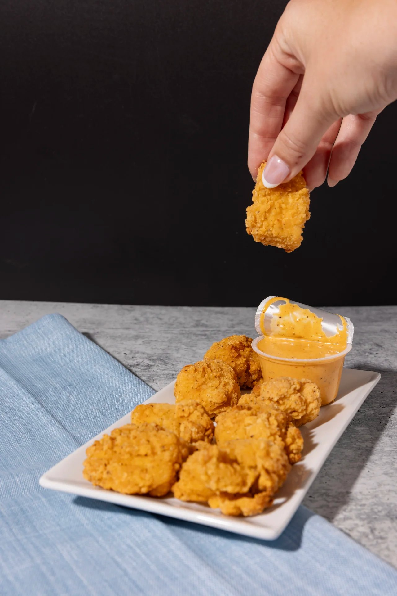 Hand holding chicken nugget above a plate with dipping sauce